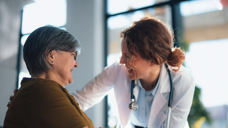 A compassionate doctor leans toward a smiling senior woman in a hospital office, offering care and reassurance during a medical consultation focused on health, trust and patient-centered care, m3 mi, Why rare disease strategy needs a connected view