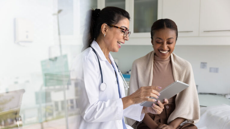 Happy senior practitioner woman and cheerful young African patient woman watching examination medical report on tablet computer, using digital technology, laughing